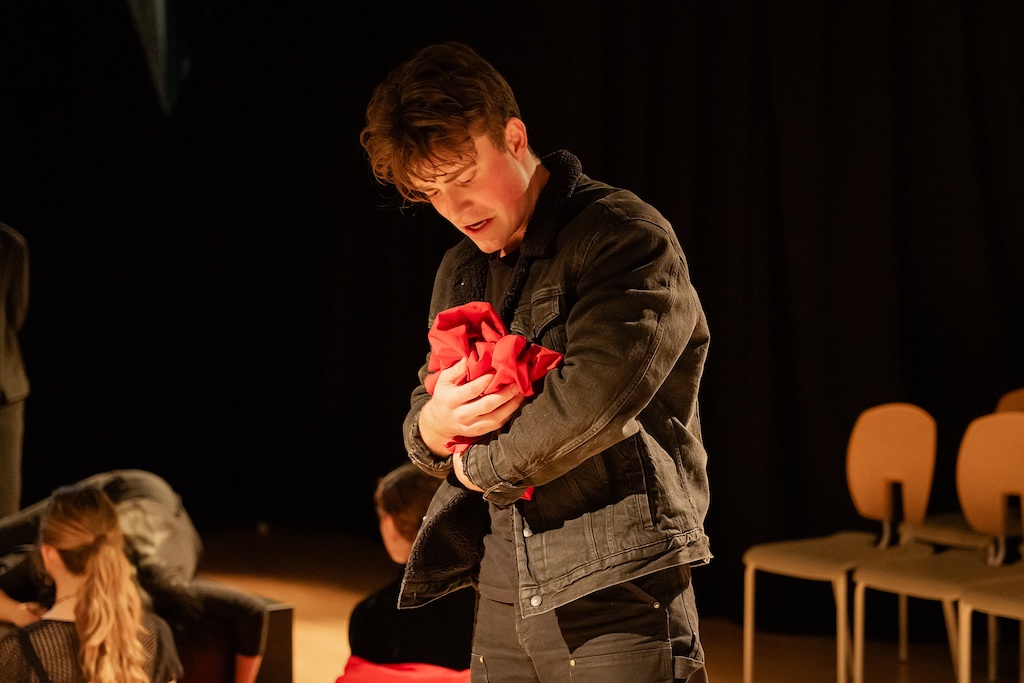 An actor stands on stage in mid-performance, looking down at the bright red cape he is holding.